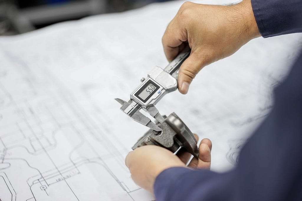 a man in front of device schematics measures a metal components thickness with a caliper tool.