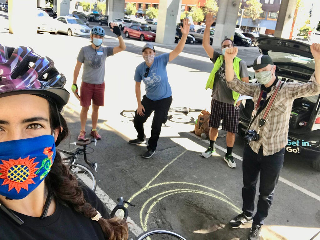 Five people with bikes stand in a parking lot, wearing masks, with hands raised in excitement after a bike education class.