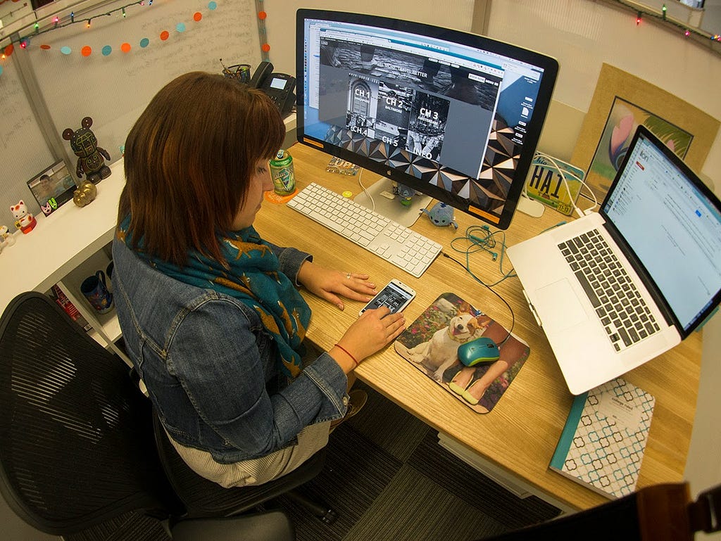 Image features the author sitting at a large wooden desk with a large monitor and laptop to the left. A black and gray eBook is featured on a large monitor and also being viewed on an Android mobile phone on the desk. On the laptop to her right is a blank screen.