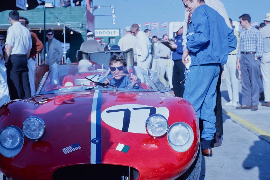 Wearing a blue racing suit and sunglasses, Denise McCluggage poses in the driver’s seat of a red racecar with the number 77 on it.