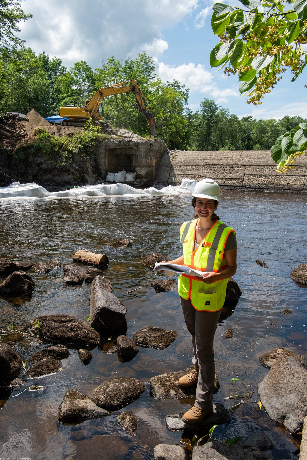 a woman in a reflective vest and hard hat stands on rocks near the river’s edge below a dam