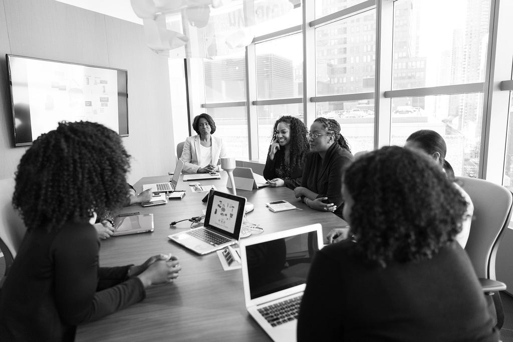 Black and white image of diverse women around a conference table with meeting items inc. laptops and notepads.