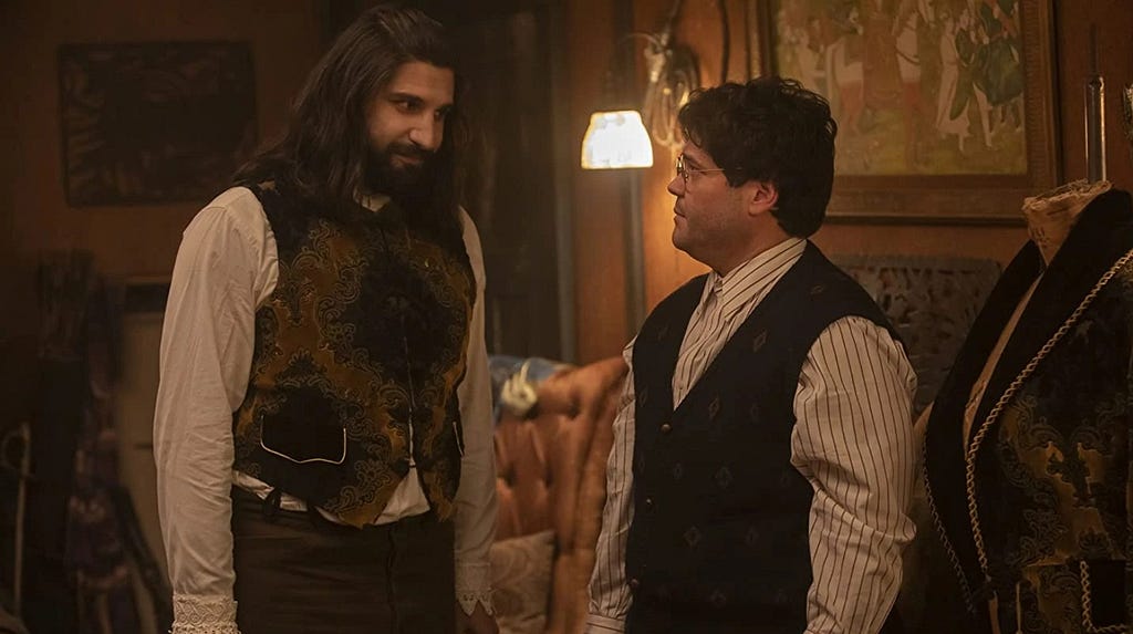 Nandor The Relentless (Kayvan Novak) in a dress shirt and short waistcoat looking at his familiar, Guillermo (Harvey Guillen) who is dressed in a striped white dress shirt with a full-length waistcoat