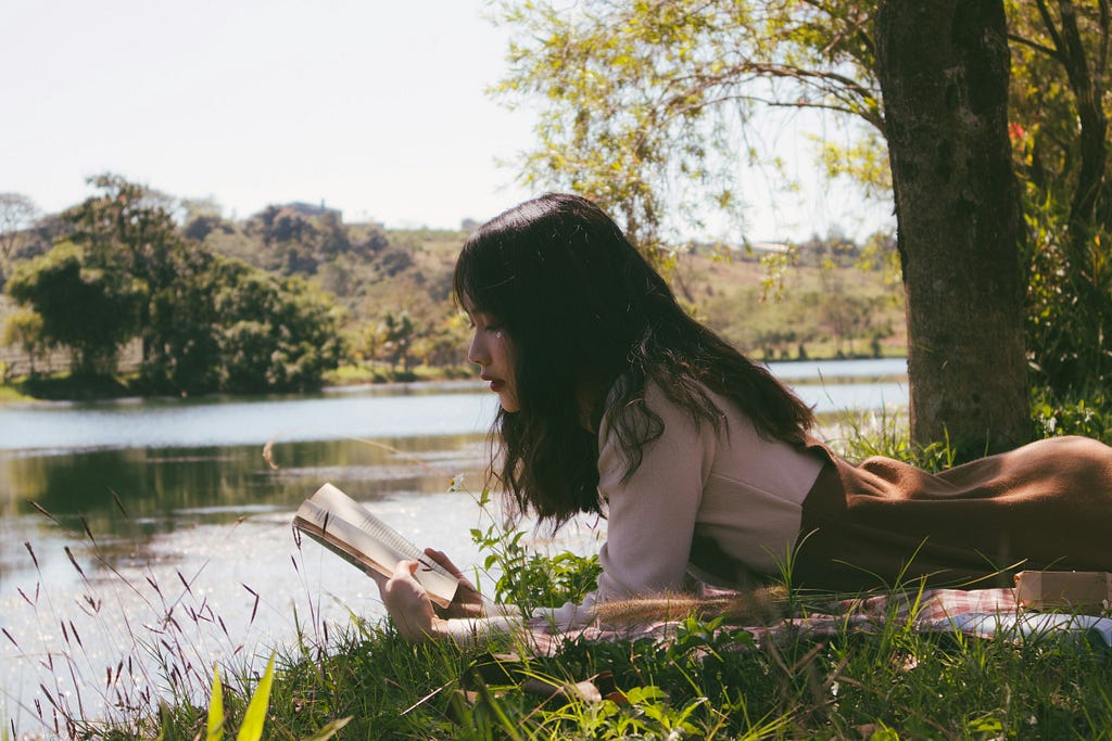 Woman reading book and lying forward on sheet on grass beside body of water during the day.E.g.