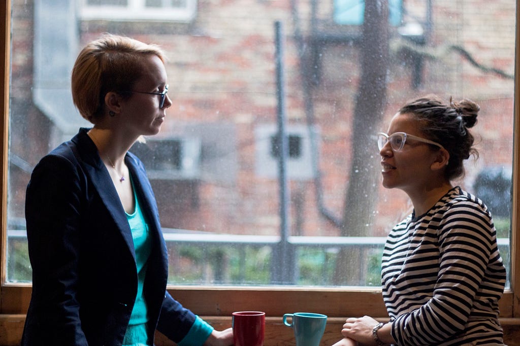 Two women conversing in front of a rainy window.