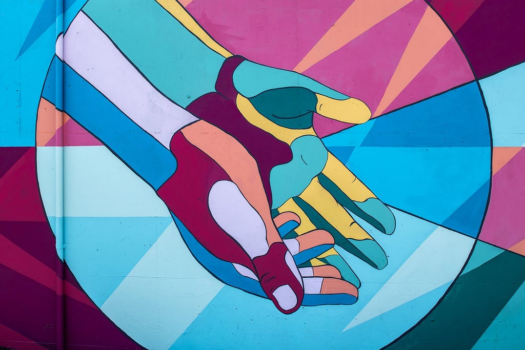 A colorful mural featuring featuring different shades of blue, pink, green, purple and yellow and forms two hands extended out is photographed.