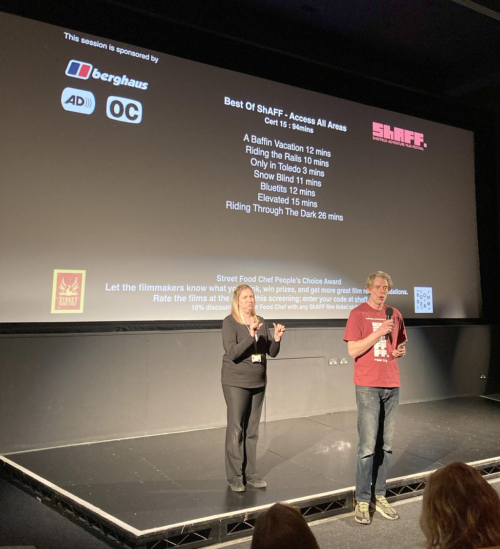 Sheffield Adventure Film Festival director Matt Heason, a white man in his late forties with blond hair wearing blue jeans and a red festival t-shirt, stands on stage at a cinema with a sign language interpreter beside him signing. She is white, in her late thirties and wearing black top and trousers. On the screen are symbols for open captions and audio description and a list of films showing in the session.