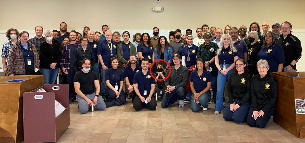 A group photo of Pittsburgh’s Department of Innovation and Performance, taken in an office lobby. Many employees wear garments with the I&P logo. A red circle marks the author crouching near the front masked.