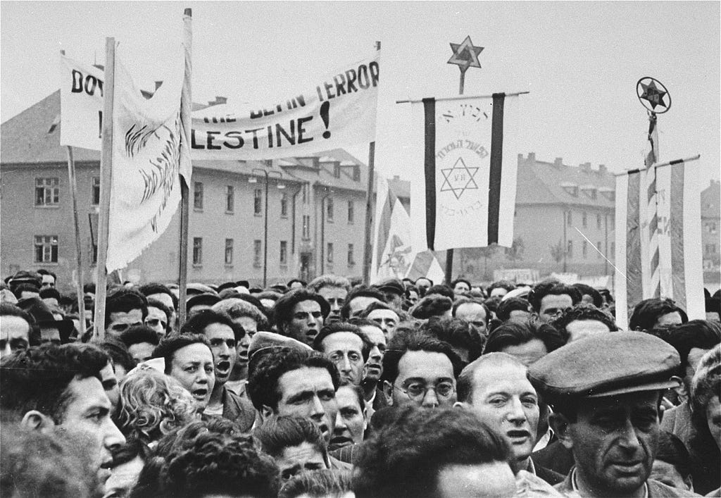 A large group of men and women crowd together in a street. A few people hold signs that contain Stars of David.