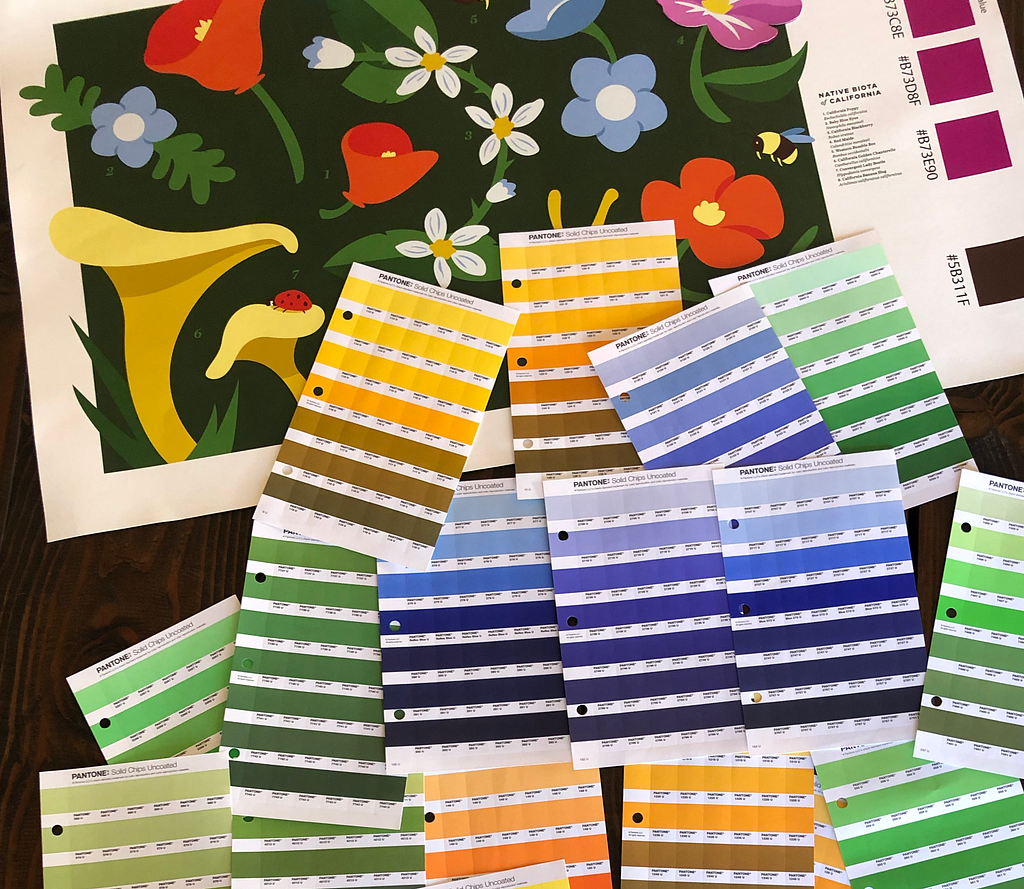 A small, paper version of the final mural design is resting on a table with approximately a dozen paint chips scattered on top of it, representing the colors that will be used to paint the mural on the wall. They are mostly blues, greens, browns, and yellows.