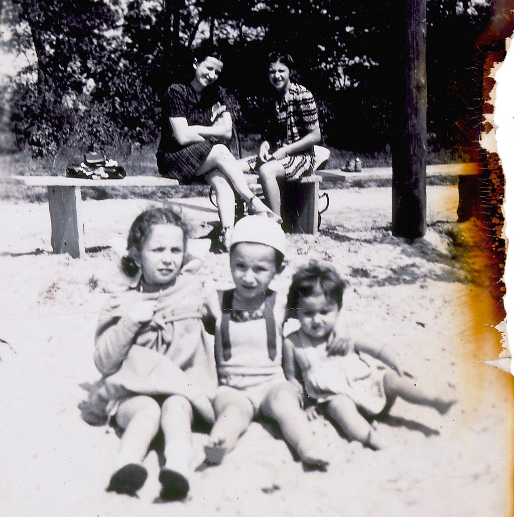 In the foreground, three young children sit in the sand with their arms around each other. In the background, two women sit on a rustic wooden bench in the sunshine, smiling and watching the children. The right edge of the photograph is browned and ragged.