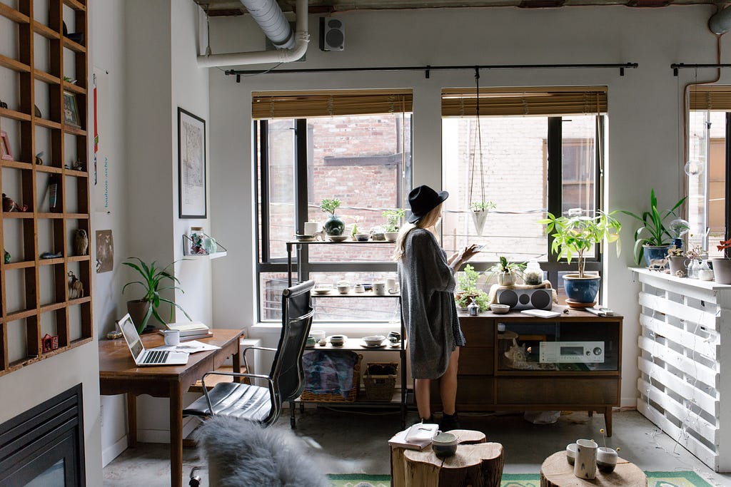 Woman in a grey dress stands in her living room, home office set up with lots of plants