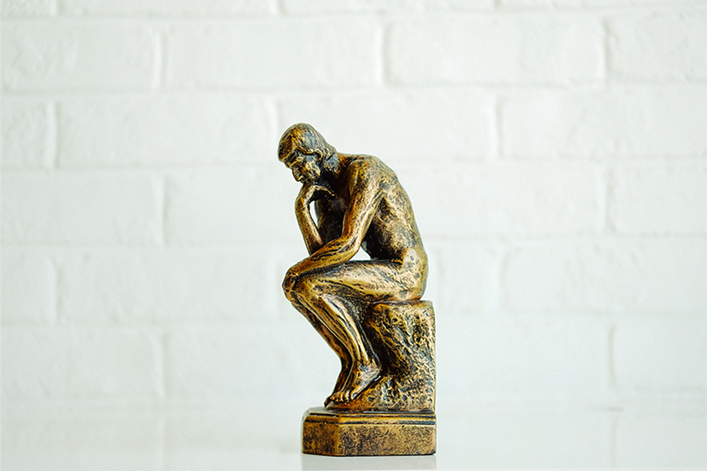 Estátua em minuatura da obra “O pensador”, dourada com tons pretos, em um fundo branco.