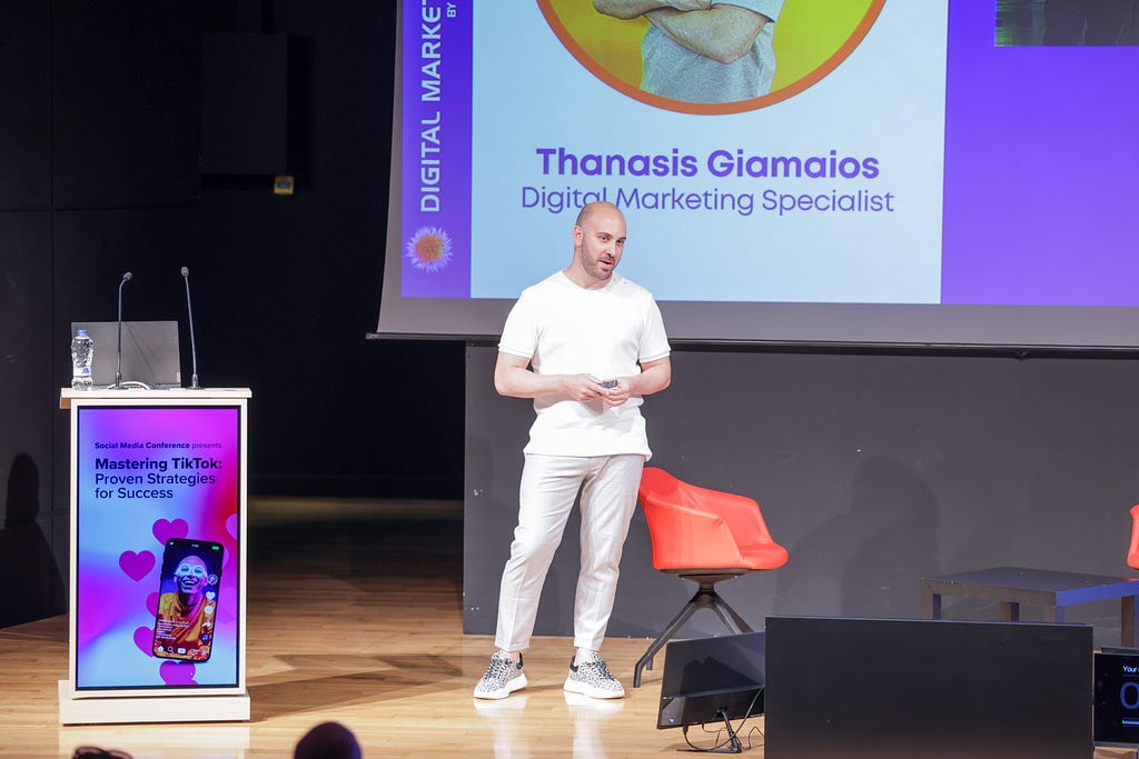 The image shows Thanasis Giamaois, a Digital Marketing Specialist, presenting on stage at the “Mastering TikTok: Proven Strategies for Success” conference. He is standing in the center, dressed in a white outfit, and holding a small device or remote in his hands. To his left, a podium displays the conference title and an image associated with TikTok. The screen behind him shows his name and title along with his photo and a vibrant background that includes the logo of the Digital Marketing event.