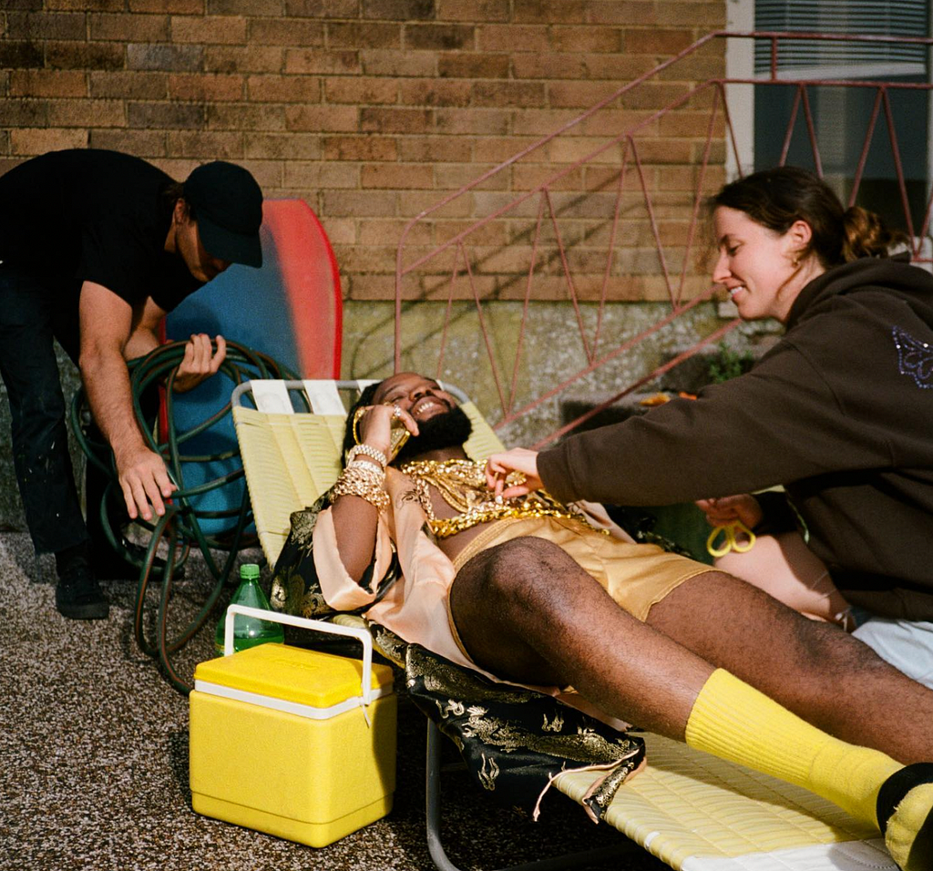 Genesis Owusu is reclining on a yellow banana lounge, outdoots, in front of what looks like a suburban house. A couple of production assistants are helping him, one of them is helping arrange the many gold chains laid across his chest. There is a small yellow Eski sitting next to the banana lounge too.