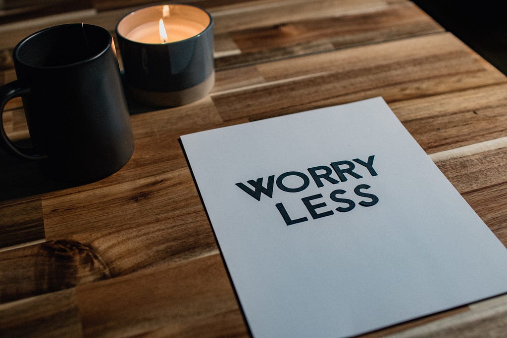 A piece of paper with the words “Worry Less” printed on it, laying on a wooden table with two black candles lit next to it.