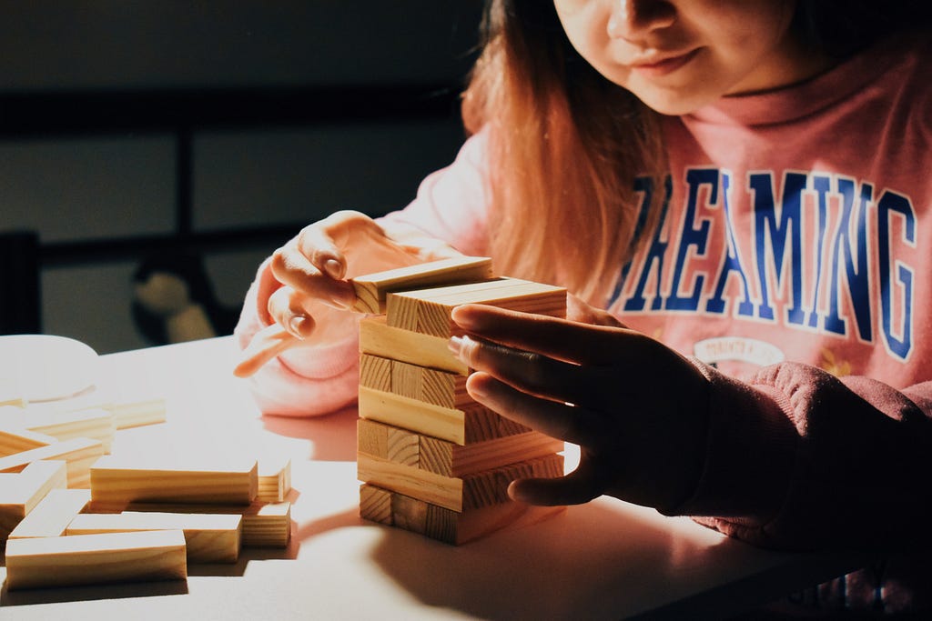 A child in a pink jumper that reads “dreaming” stacks wooden blocks on top of each other on a white desk.
