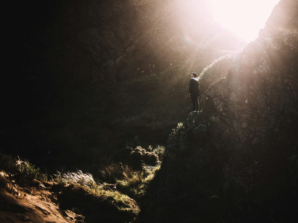 This image captures a person standing on a rocky outcrop, bathed in the warm glow of the sun peering through a narrow mountain pass. The setting is serene and isolated, evoking a sense of solitude and contemplation in nature.
