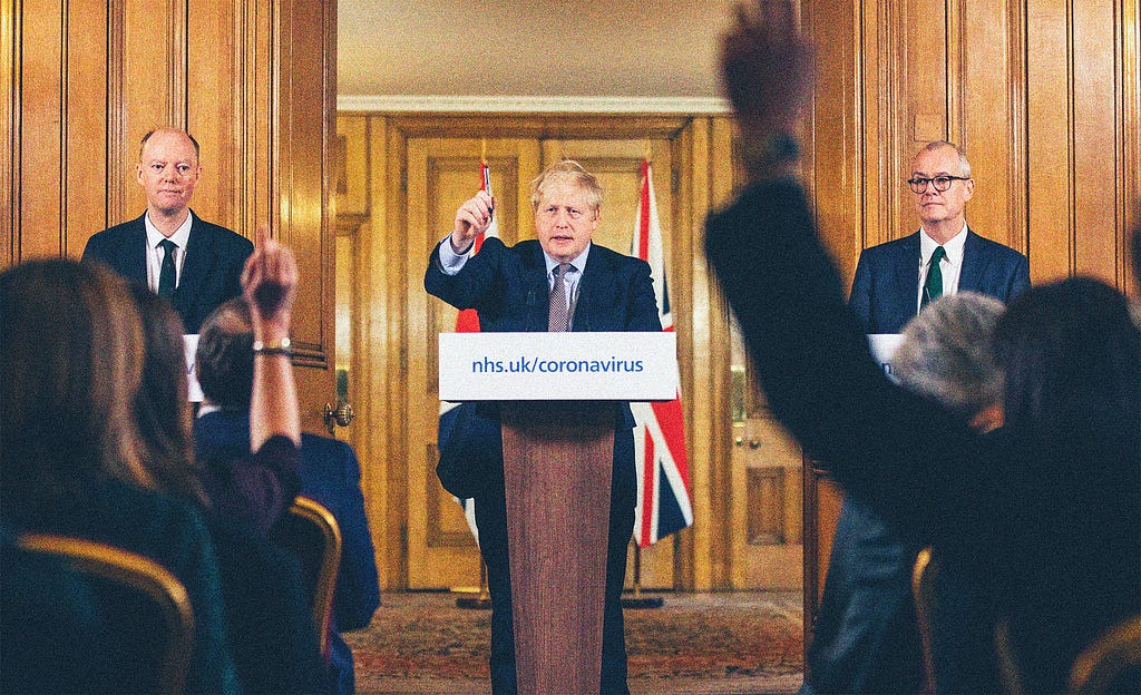 Boris Johnson standing behind a podium for a live press briefing. The podium placard reads “nhs.uk/coronavirus” in blue text.