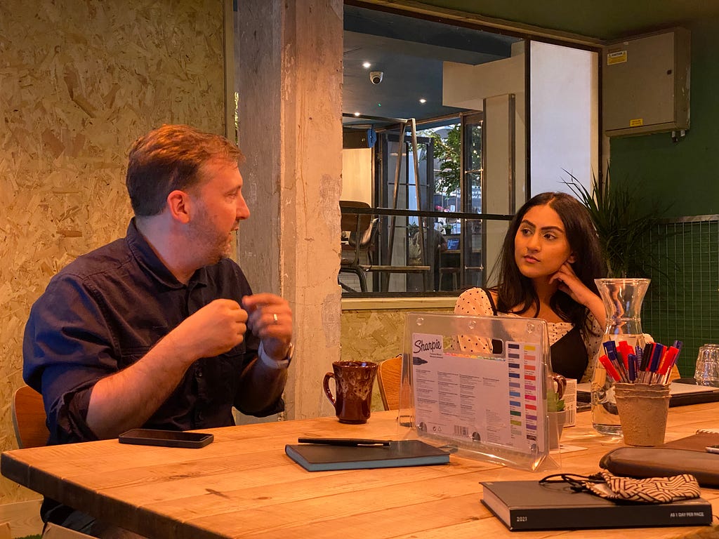 Andy (left) and Shazia (right) talking at a table, with a pot of sharpies in front of them. Credit: TBIJ