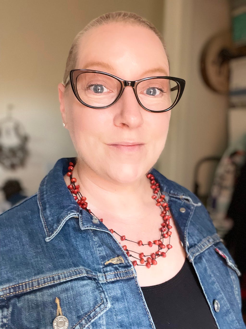 Woman with chemo hair growing out, wearing glasses, a red necklace and a denim jacket