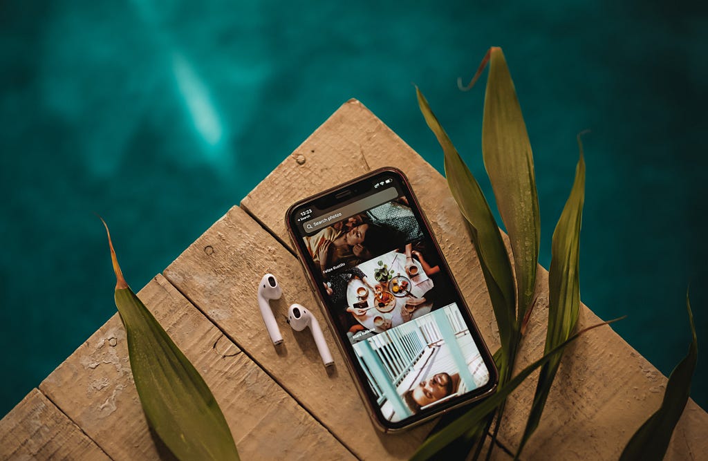 A smartphone and wireless earphones are laid on the edge of a wooden table. The background is a marine blue, the table is presumably near a body of water such as a pool.