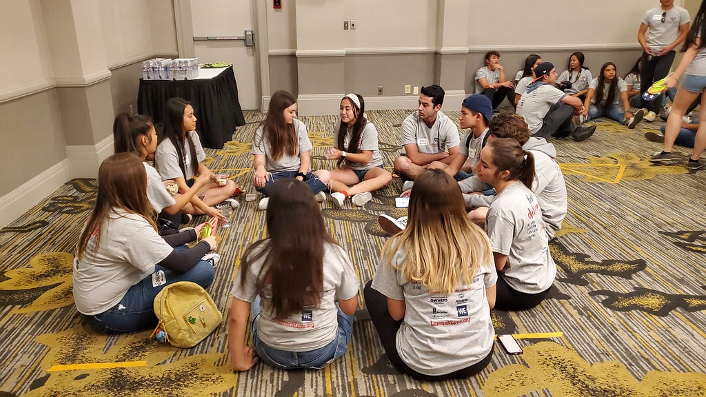 A group of Linens N Love volunteers all wearing white shirts and sitting in a circle on the floor. They are discussing their changemaking journeys.