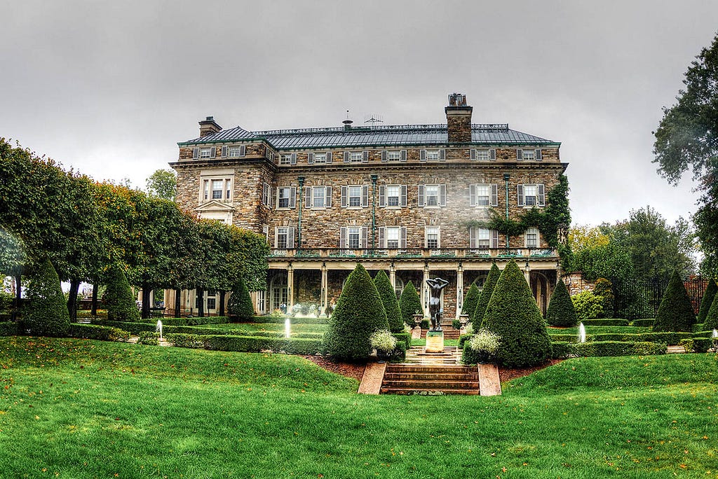 A grand stone mansion with a symmetrical facade and a red tile roof. The house is surrounded by a well-maintained lawn and mature trees.