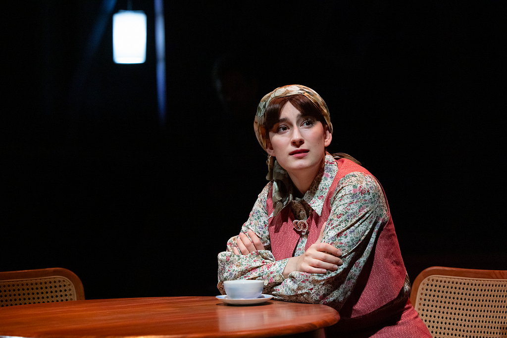 A woman wearing a kerchief over her hair sits at a table with a cup of tea and looks out wistfully.