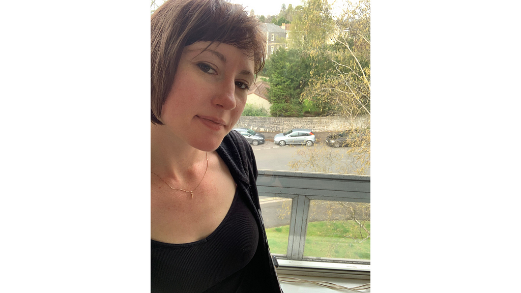 Selfie of Elizabeth standing beside a window overlooking trees and buildings.