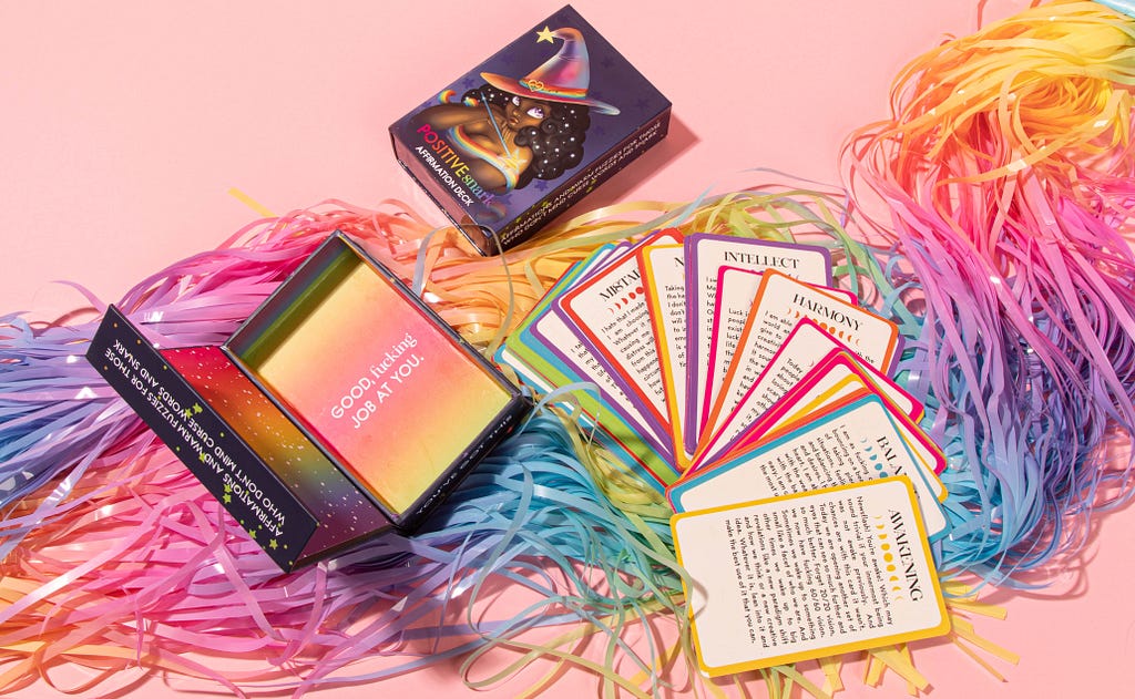 A deck of affirmation cards sits on rainbow tassles laid out. The cards are rainbow and the box says “Good f*cking job at you” on the inside.