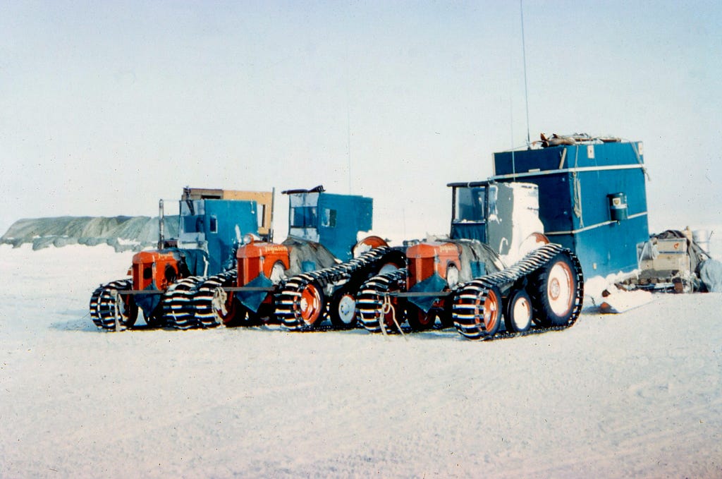 3 converted farm tractors with tracks over their wheels. They are parked in the snow in Antarctica. one is towing a cabin called a caboose that the drivers used for shelter.