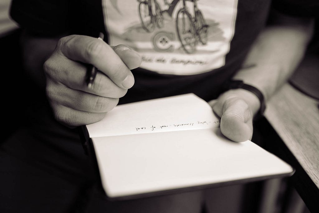 The picture shows a man holding a book on which he writes with a pen. It is a black and white image. The pages are mostly blank, but he has written one sentence.