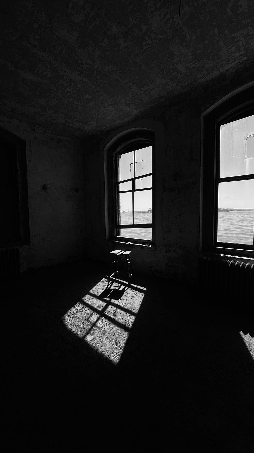 Black and white photo of a chair by a window in a room at the abandoned Ellis Island Immigrant Hospital by @1234