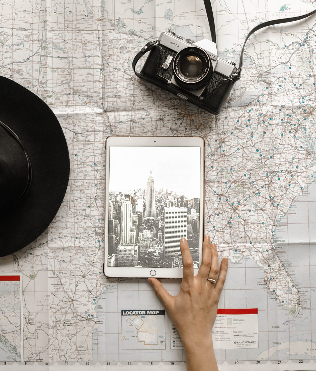 A tablet showing an image of New York, sat on a map. A hat and camera lay on the table too.