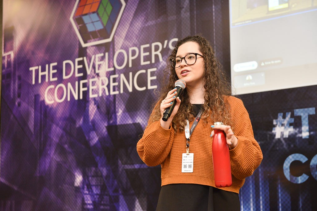 Ao fundo o painel no palco principal do TDC, tem o logo que é um cubo mágico colorido seguido do nome do evento: the developer’s conference. Em cima do palco estou segurando com uma mão o microfone enquanto explico. Sou uma mulher branca, de cabelos longos castanhos e cacheados. Estou usando um blusão de lã laranja.