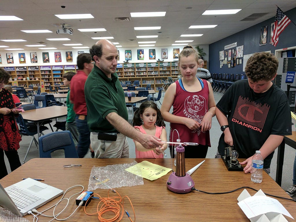 The author demonstrates wireless energy produced by a Tesla Coil to middle school students by illuminating a neon tube nearby.