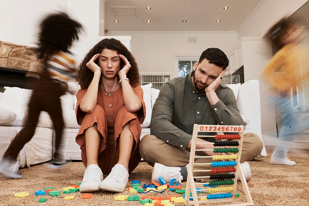 Overwhelmed parents on the floor, children running with toys in a chaotic living room, capturing family stress and motion blur.