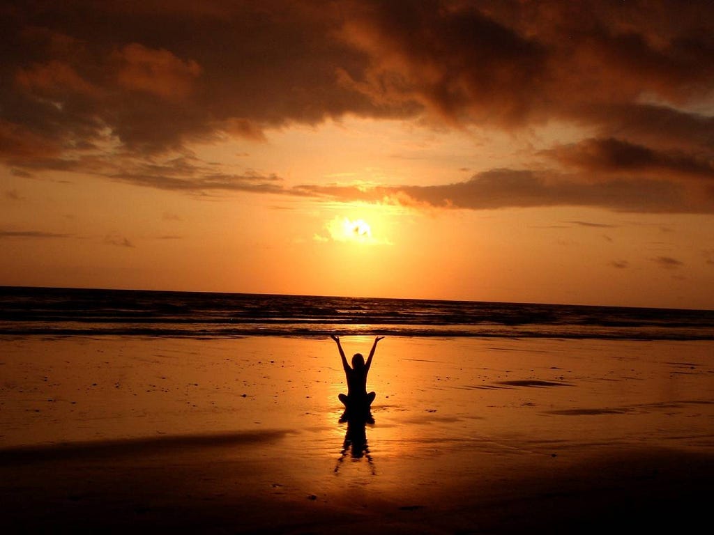 person sitting on a beach with arms raised at sunset