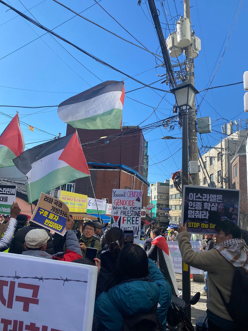 Author photo of a Palestine protest in Itaewon Seoul. Three Palestinian flags wave in the bright sunlight against a blue sky and there are posters in Korean and English.