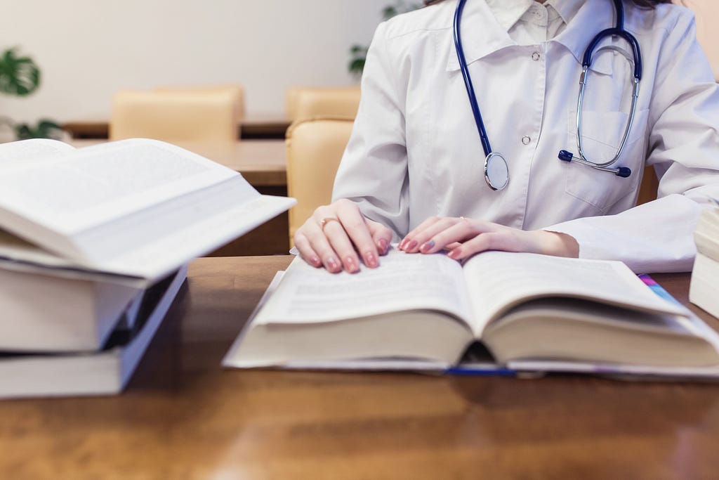 A student with a stethoscope around their neck studies a textbook that is open on the desk in front of them.