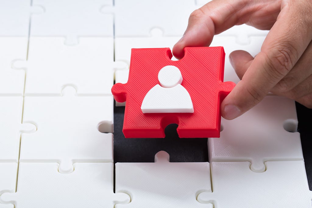 Hand putting red puzzle piece with a user icon on it as the last piece into a bland white puzzle.