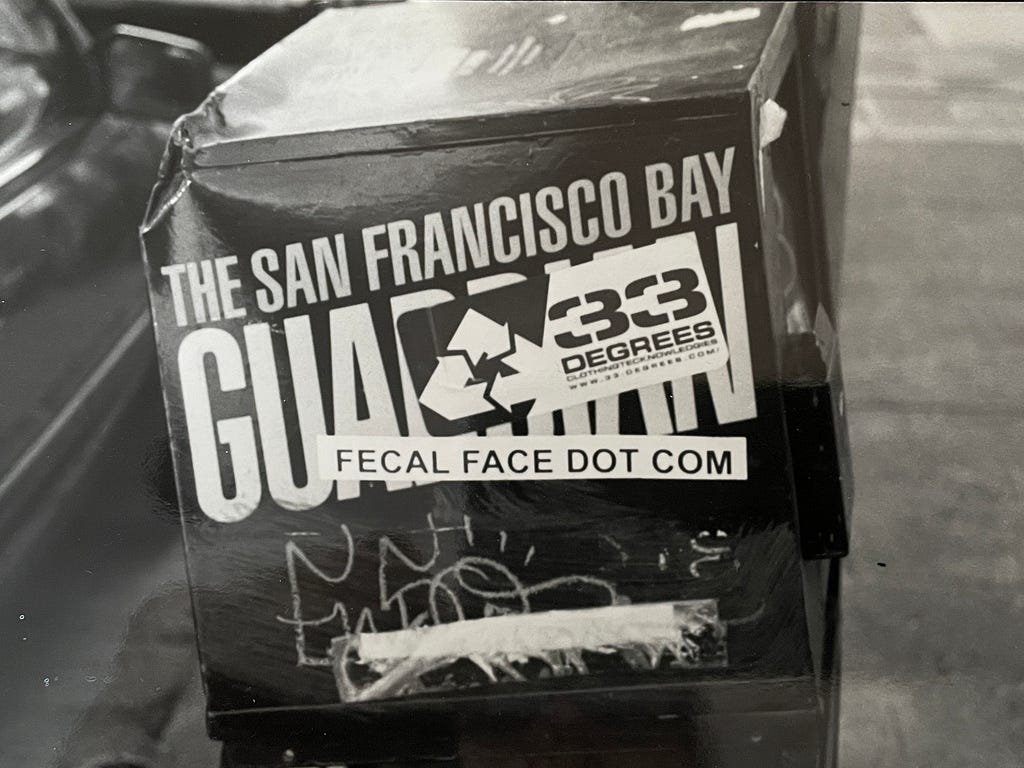Photo of a San Francisco Bay Guardian paper rack with a sticker on the side that says fecal face dot com.