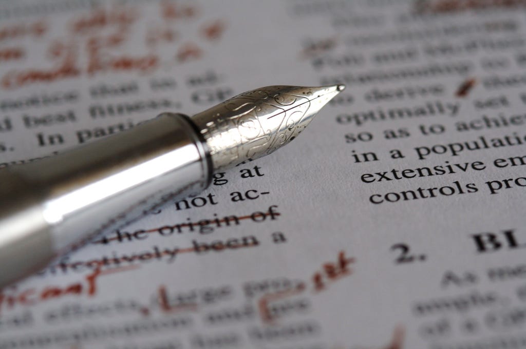 Photo shows closeup of a pen sitting on a printed document covered in edits written in red pen.