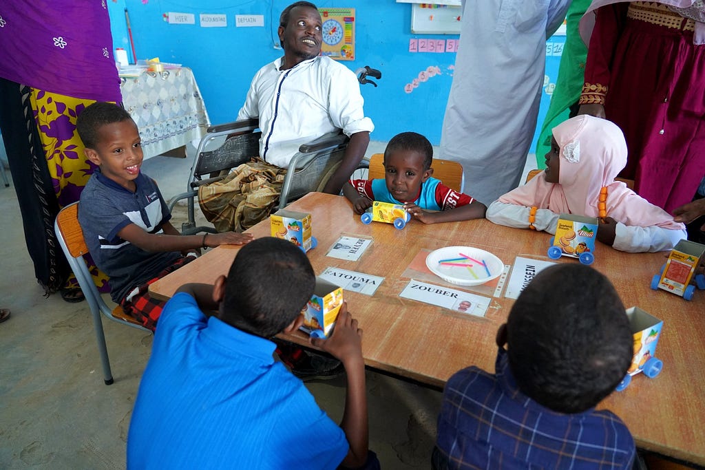 A parent who uses a wheelchair gazes up towards one of the teachers in a classroom. The parent sits at a table with five preschool students.