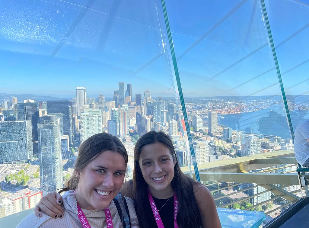 Jacqueline and Amelie at T-Mobile Changemaker Challenge Spaceneedle event. They are inside of the Space Needle with a beautiful view of the city behind them.