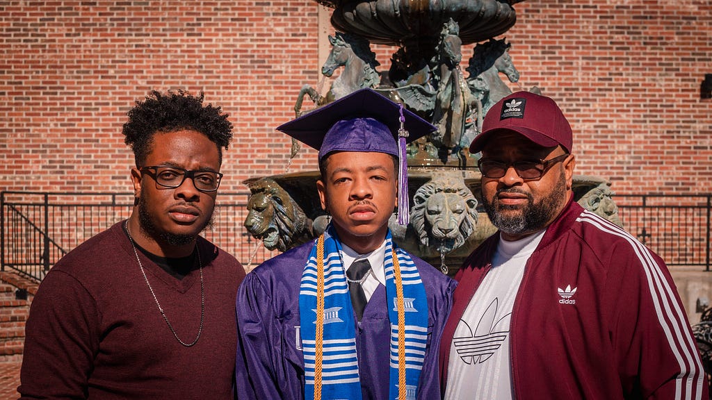 2 young black men and their father. One of the young men is in graduation attire.
