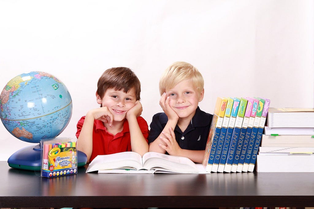 Two children sitting and smiling with textbooks, a globe, and some markers