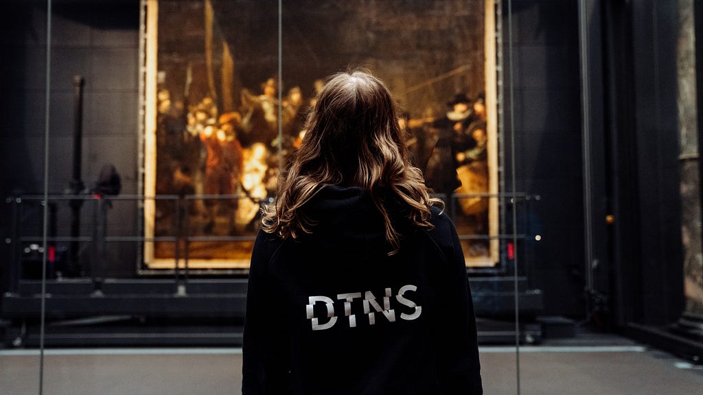 The picture features a woman’s back. She wears a black hoodie that features the letters DTNS on the back in white. She is looking at a Rembrandt painting.