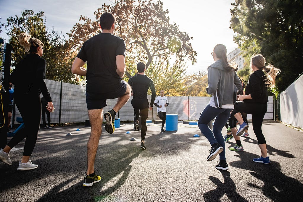 A group of people exercising in the middle of a park, on a paved surface, with an instructor guiding them at the front.